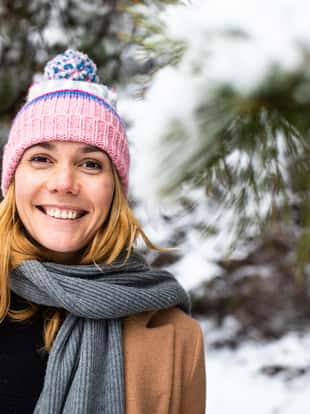 A pretty young blonde in warm clothes is smiling at the camera while standing in a snowy forest