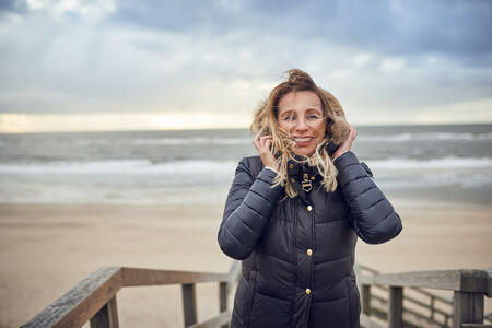 Middle-aged woman braving a cold winter day at the seaside standing on a wooden deck overlooking the beach on a breezy day smiling happily at the camera