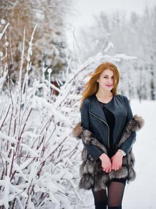 Red haired girl in fur coat walking at winter snowy park.