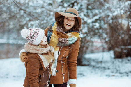 smiling stylish mother and daughter in hats and sheepskin coats with mittens outdoors in the city park in winter.