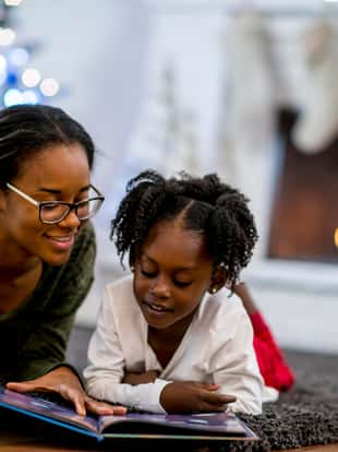 A mother and daughter of African descent are in indoors in their living room during Christmas time. They are wearing warm, comfortable clothes. They are laying on the carpet and reading a storybook together.