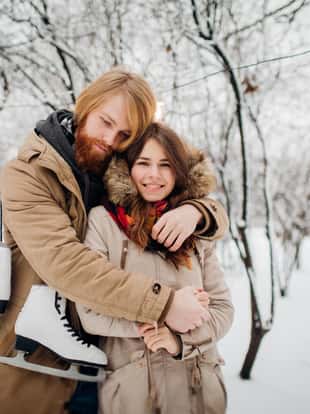 Winter and date. Young couple in love with man and woman in the winter against background of snow covered trees in park are embrace. A guy with long hair and beard keeps skates and loves girlfriend.