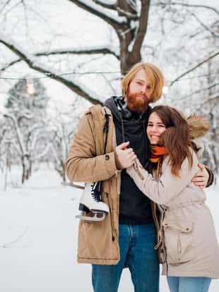 Winter and date. Young couple in love with man and woman in the winter against background of snow covered trees in park are embrace. A guy with long hair and beard keeps skates and loves girlfriend.