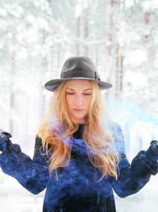 Portrait of a young blonde fashion model in the snowy forest, holding coloured smoke fountains.