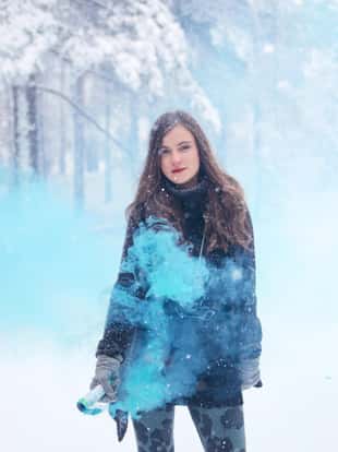 Portrait of a brunette fashion model in warm winter clothing, holding a colourful smoke fountain, walking in the snowy forest.