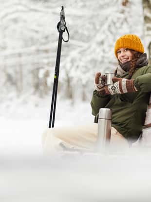 Side view portrait of active young woman smiling at camera while enjoying hot cocoa sitting on bench in beautiful winter forest, copy space