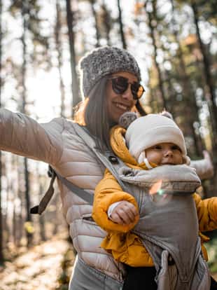 Beautiful smiling young mother carrying her baby in baby carrier and walking through forest on sunny day