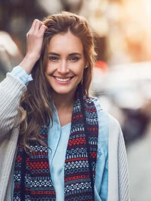 Happy young adult woman smiling with teeth smile outdoors and walking on city street at sunset time wearing winter clothes and knitted scarf.