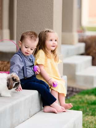 Well-dressed little boy and girl sitting on the front porch, barefoot, with easter baskets, holding eggs, and looking at the bunny.