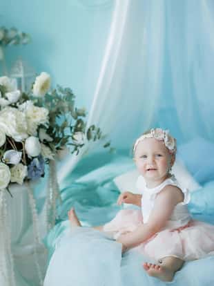 Little baby 1 year from birth sitting on a bed or on a chair in a pale blue children's room.