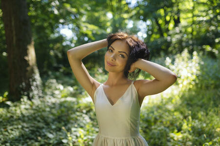 Close-up of a young woman enjoying the sun, fresh air and freedom. She's cute and smiling, and wears white dress in hot summer afternoon. She holds hands in her hair and looking at the camera. Shallow DOF. There are trees and lush foliage in the background. Whole scene is illuminated by sunlight from behind. Low depth of field and blurred background.