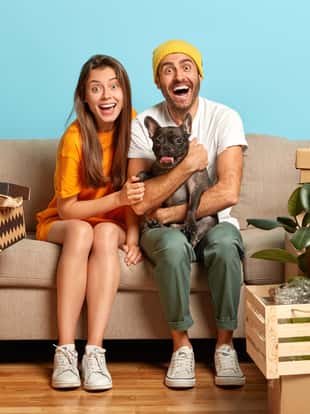 Photo of overjoyed married family couple rest on sofa during moving day, moved into new apartment, play with black pedigree bulldog, many packages with household items around. Two renters in abode