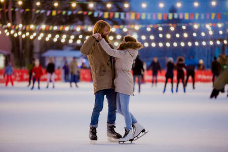 Theme ice skating rink and loving couple. meeting young, stylish people ride by hand in crowd on city skating rink lit by light bulbs and lights. Ice skating in winter for Christmas on ice arena.