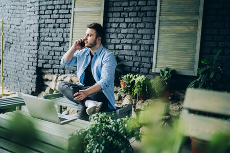 From above of male business owner in casual clothing sitting with crossed legs and looking away while talking on mobile phone at outdoors terrace of cafe