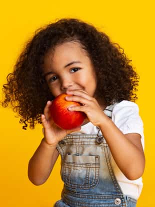 Yummy. Little afro girl eating red apple and looking at camera over yellow background