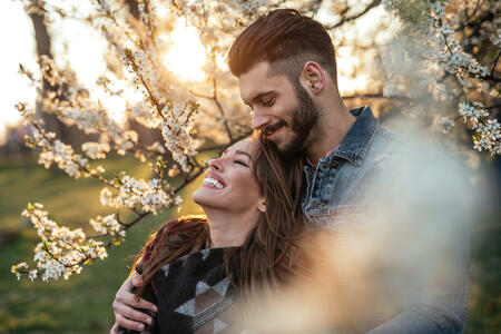 Photo of a couple enjoying in the park at sunset. The man is embracing his girlfriend from behind. They are both happy.