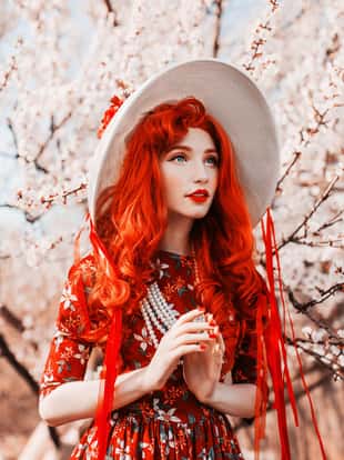 Young handsome redhead girl in spring flower garden. Woman in vintage hat and  retro dress on nature. Beautiful spring cherry tree. Handsome redhead model in retro hat. Vanilla flower dress. Red nails