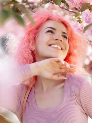 Close-up portrait of bright young woman with curly pink hair and sunglasses near the blossoming spring tree