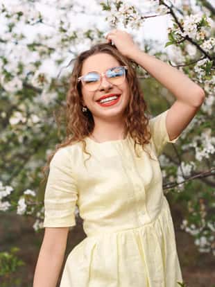 Happy stylish lady in dress and glasses smiling and touching wavy hair while standing near tree with white flowers on sunny day in spring garden