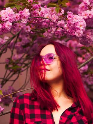 Gorgeous brightful model with burgundy colored hair covering half of the face. Branches of sakura tree fully in bloom on background