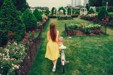 a cute and stylish girl in a yellow dress and long hair rides on a bicycle with a basket of flowers in the sunny summer garden