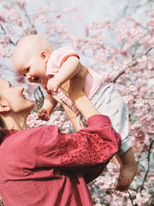 Loving mother and baby girl on background blooming pink flowers of apple tree at springtime. Beautiful woman with daughter among garden flowers outdoors. Family on nature in Arboretum, Slovenia