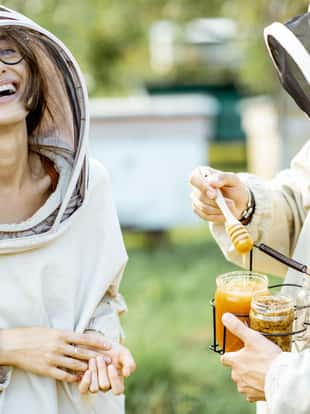 Portrait of a cheerful man and woman beekepers in protective uniform standing together with honey in the jar, tasting fresh product on the apiary outdoors