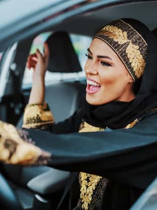 Cheerful gorgeous positive muslim woman in traditional wear driving her new car, listening music and singing.