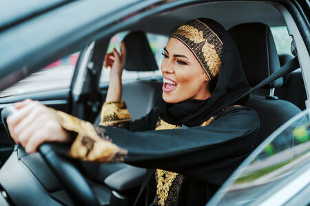 Cheerful gorgeous positive muslim woman in traditional wear driving her new car, listening music and singing.