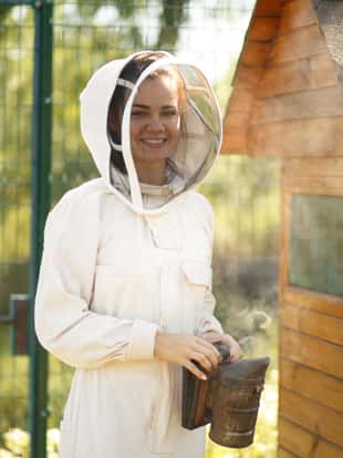 Beekeeper girl in white suit posing near a clue with a bee smoker.