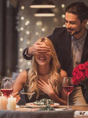 Handsome elegant man is holding roses and covering his girlfriend's eyes while making a surprise in restaurant, both are smiling