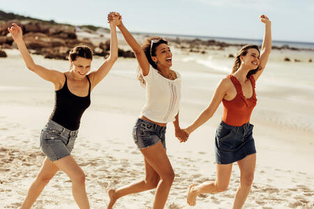 Happy women friends walking together on beach holding hands. Three young women enjoying and dancing together on a beach vacation.
