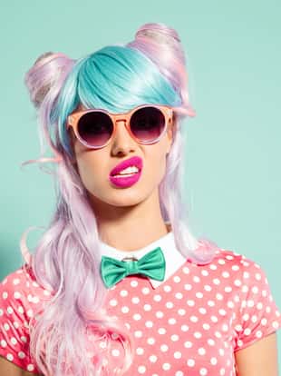 Portrait of manga style blue-pink hair girl wearing sunglasses and pink polka dot dress with collar and bow tie. Standing against turquoise background, looking at camera. Studio shot, one person.