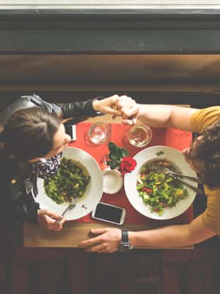 Above view of young romantic couple holding hands during their date in the restaurant. There are plates with food, glasses with drinks, rose and mobile phones on the table.