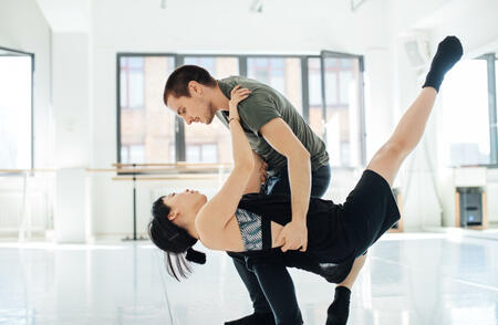 Side view of young male dancer holding female on dance floor. Multi-ethnic man and woman are practicing ballet dance at studio. They are performing against window.