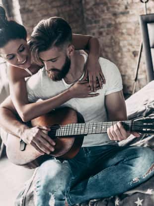 Handsome young bearded man playing guitar while attractive woman embracing him and smiling