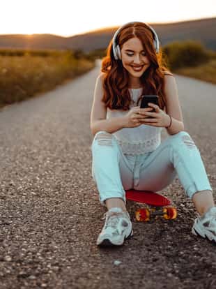 Smiling teenage girl sitting at the skateboard, using headphones and a smartphone in the street on a sunny summer day