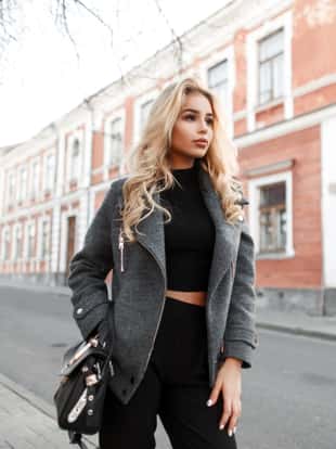 Pretty young woman model with a stylish black handbag in a fashion jacket walking on the street