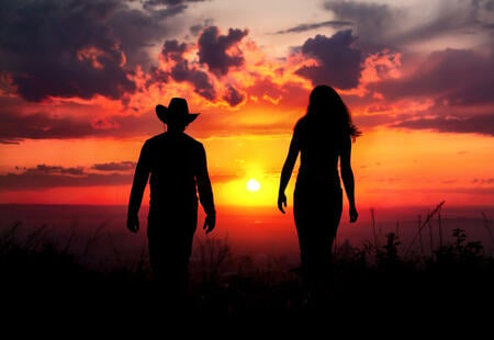 Young couple silhouette walking outdoors at sunset dramatic sky background. Man in cowboy hat and woman nearby