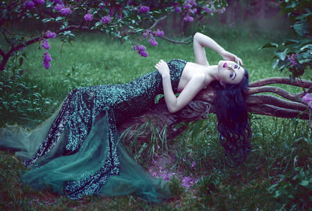 pretty slim girl with dark hair in a long emerald green dress with bare shoulders lying on a branch of a flowering lilac