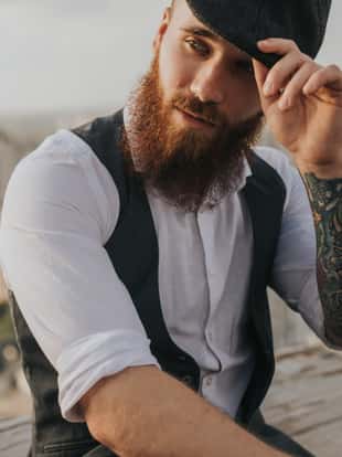 Trendy bearded man with tattooed arm looking away and adjusting retro hat while resting on rooftop on blurred background of city