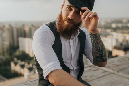 Trendy bearded man with tattooed arm looking away and adjusting retro hat while resting on rooftop on blurred background of city