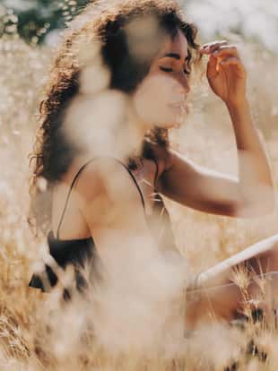 Beautiful caucasian woman with long dark curly hair is looking aside while sitting amidst the field on a blurred background. A model look female in long black dress is relaxing outdoors on a sunny day