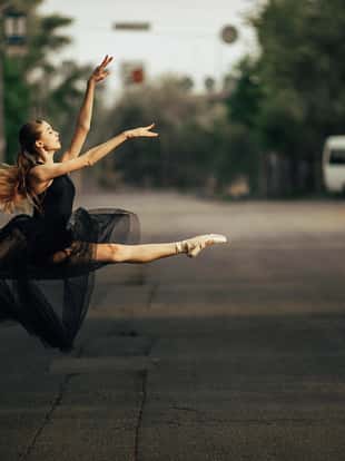 Ballerina jumping in twine pose in a black transparent dress against the background of city street.