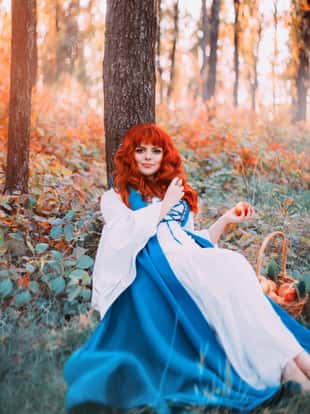cute Bavarian beauty under tree in morning light forest with basket of apples, holds red fruits in her hands, pretty girl with orange hair and snub nose and pink tender skin, sweet princess resting.