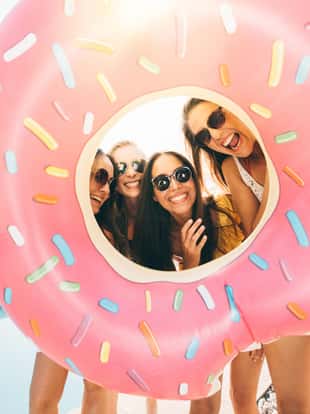Four girls holding a pink pool inflatable around their faces like a frame while laughing together and standing outdoors next to a pool