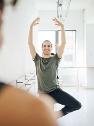 Smiling male dancer practicing ballet with arms raised. Young performer rehearsing by barre at dance studio. They are practicing at ballet studio.