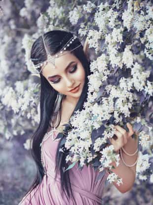 Portrait of an elf in a blooming garden. A girl with long ears touches the flowers. She is dressed in a purple dress with pearls.