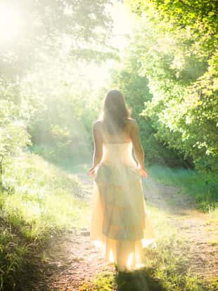 Beautiful young woman wearing elegant white dress walking on a forest path with rays of sunlight beaming through the leaves of the trees