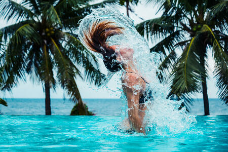 A sexy young woman is doing a hairflip and creating splashes in a swimming pool with palm trees and the ocean in the background